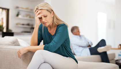 Woman sitting on edge of couch with head in hands and husband sitting in background.